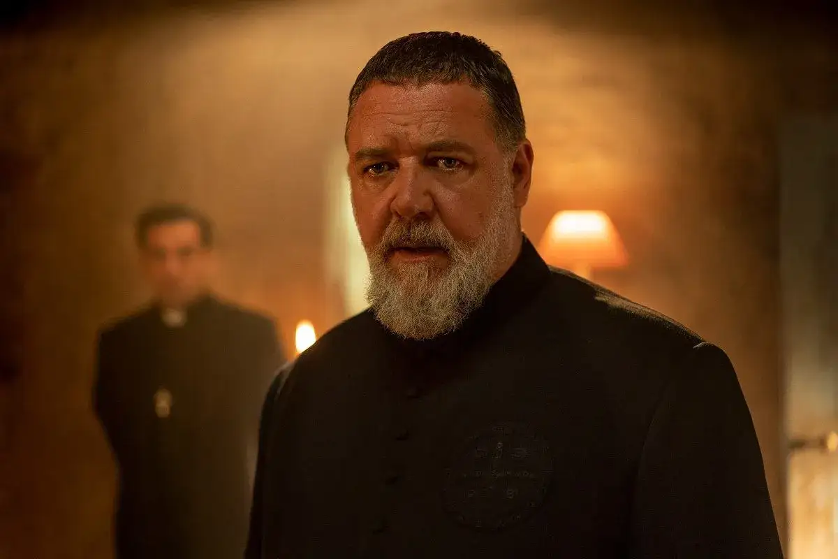 Russell Crowe, kt&oacute;ry grał papieża, w czarnym stroju duchownym. W tle inny duchowny.
