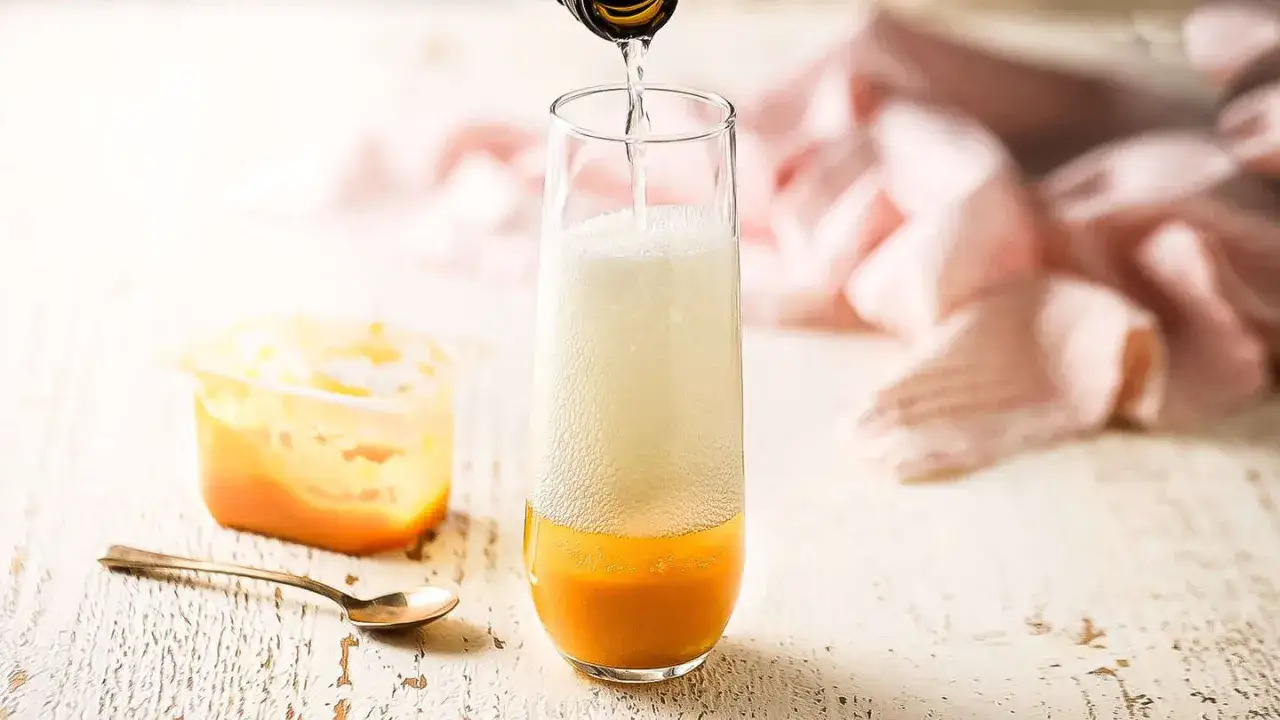 Przygotowanie drinka martini bellini: nalewanie prosecco do kieliszka z brzoskwiniowym puree. Obok pojemnik z puree i łyżeczka.