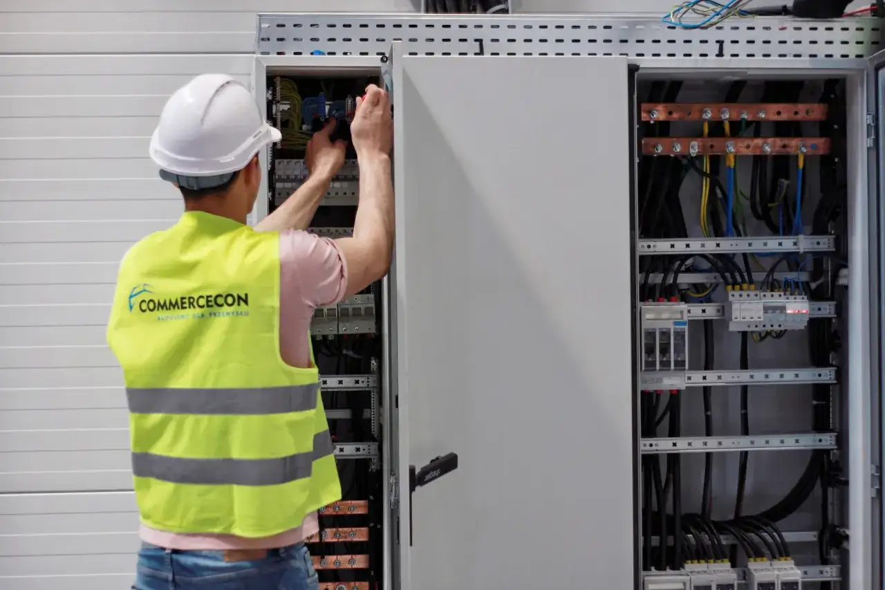 Elektryk w kamizelce odblaskowej pracuje przy rozdzielnicy, dbając o efektywność i bezpieczeństwo instalacji, co wpływa na cenę prądu przemysłowego.