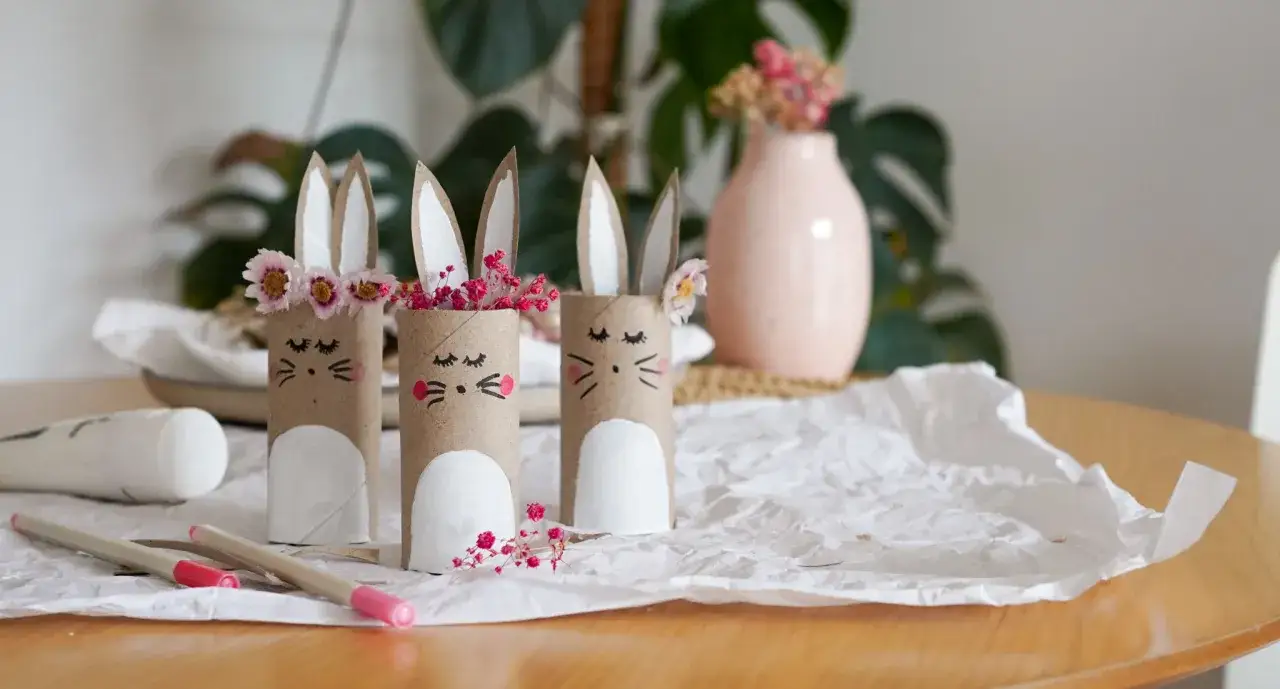 Drei s&uuml;&szlig;e Hasen aus Klopapierrollen, dekoriert mit Blumen und Stiften daneben.