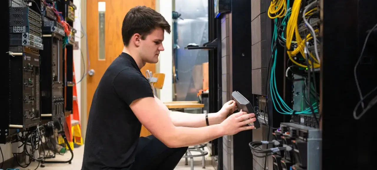 Ein junger Mann bei seiner **Netzwerkadministrator Ausbildung** installiert ein Gerät in einem Server-Rack, umgeben von Kabeln und Technik.
