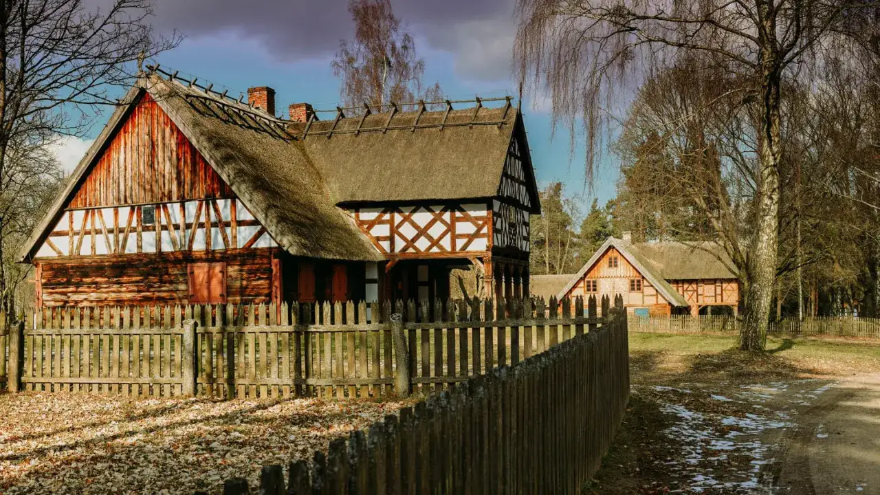 Drewniane chaty ze słomianymi dachami, typowe dla skansenów w Polsce, stoją za płotem.