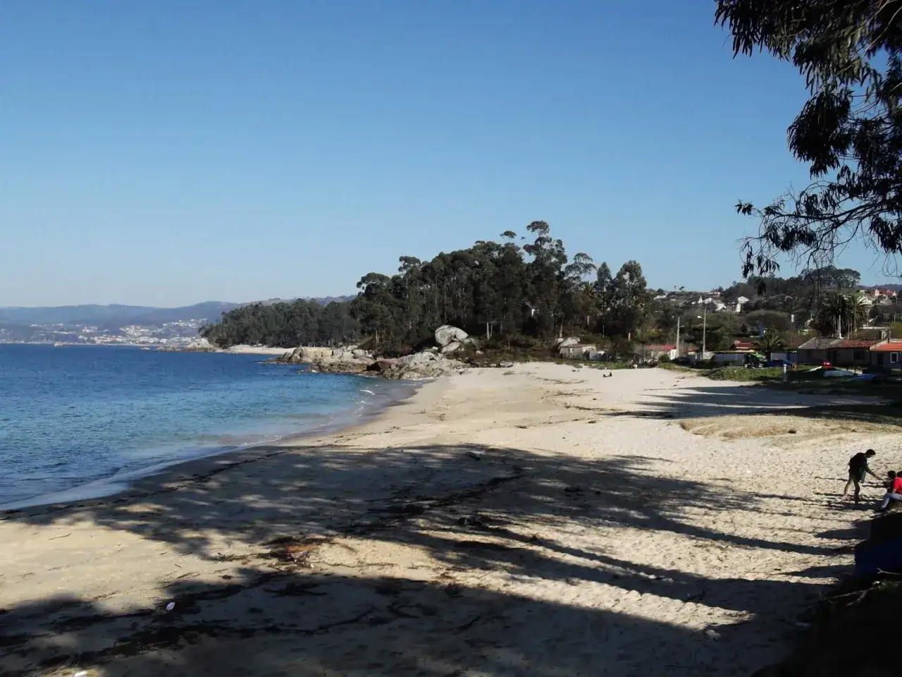 La **playa Mourisca** se extiende bajo un cielo azul, con aguas tranquilas y arena dorada. Árboles frondosos bordean la costa, y algunas casas se asoman entre la vegetación.