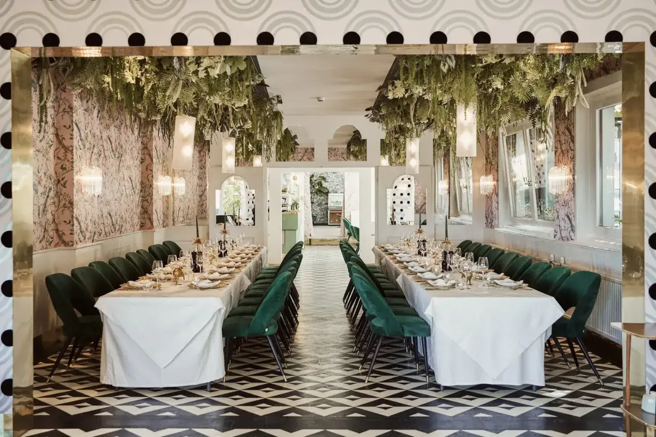 Ein Festsaal mit zwei langen Tafeln, geschm&uuml;ckt mit &uuml;ppigem Gr&uuml;n von der Decke. Gr&uuml;ne Samtst&uuml;hle s&auml;umen die Tische, bereit f&uuml;r eine Hochzeit mal anders feiern.