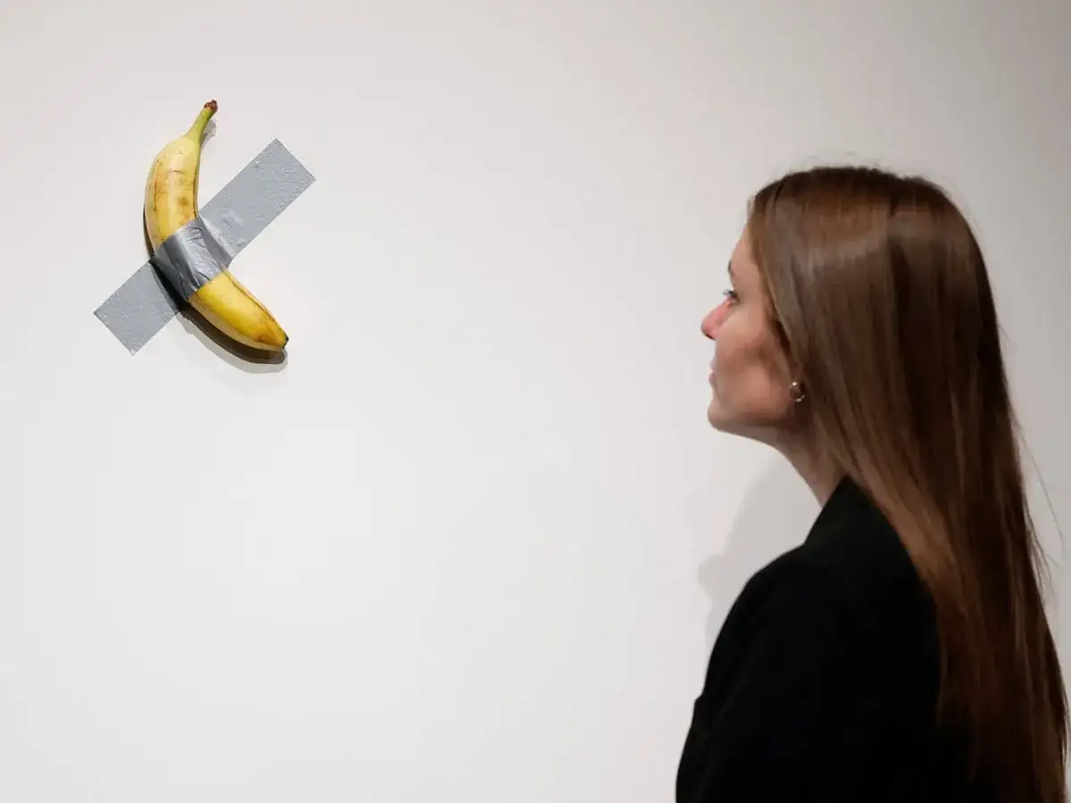 A woman observes Maurizio Cattelan's infamous banana artwork, taped to a white wall.