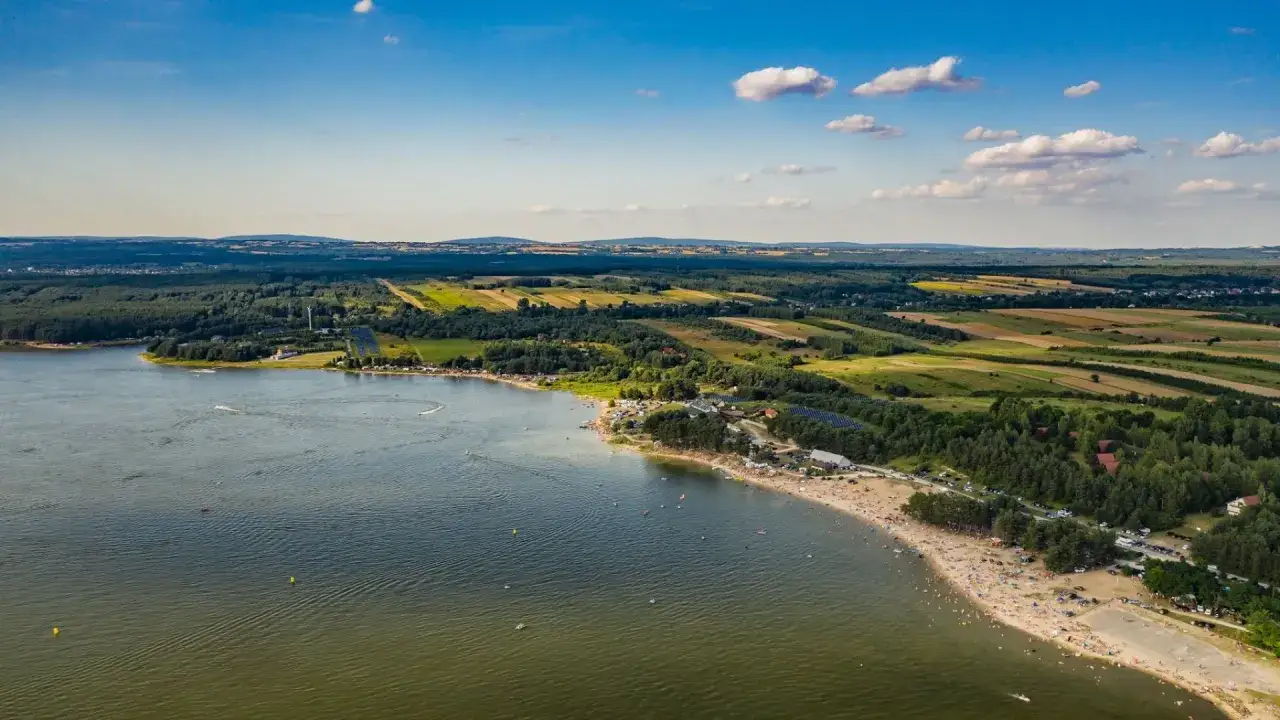 Letnie słońce nad jeziorem Chańcza, pełnym plażowiczów i wodnych atrakcji. W tle zielone lasy i malownicze pola.