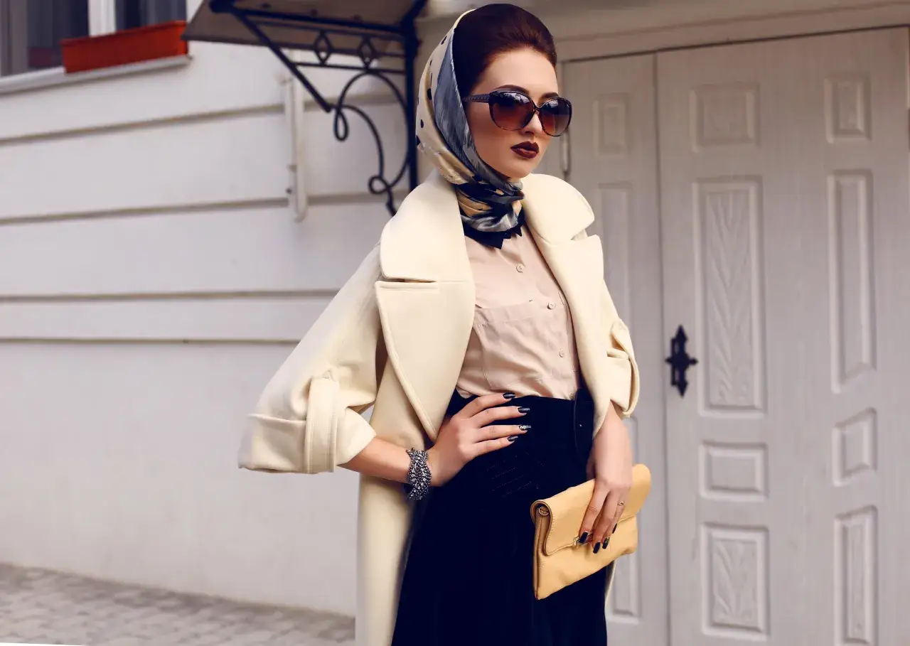 Elegancka kobieta w beżowym płaszczu, okularach i apaszce pod szyję, trzymająca żółtą kopertówkę.
