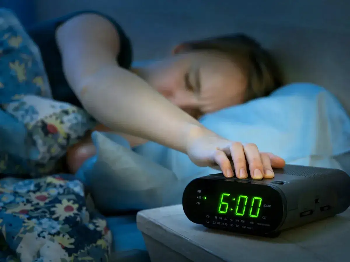 A person's hand reaches to silence an alarm clock showing 6:00 AM, questioning if snoozing makes you more tired.