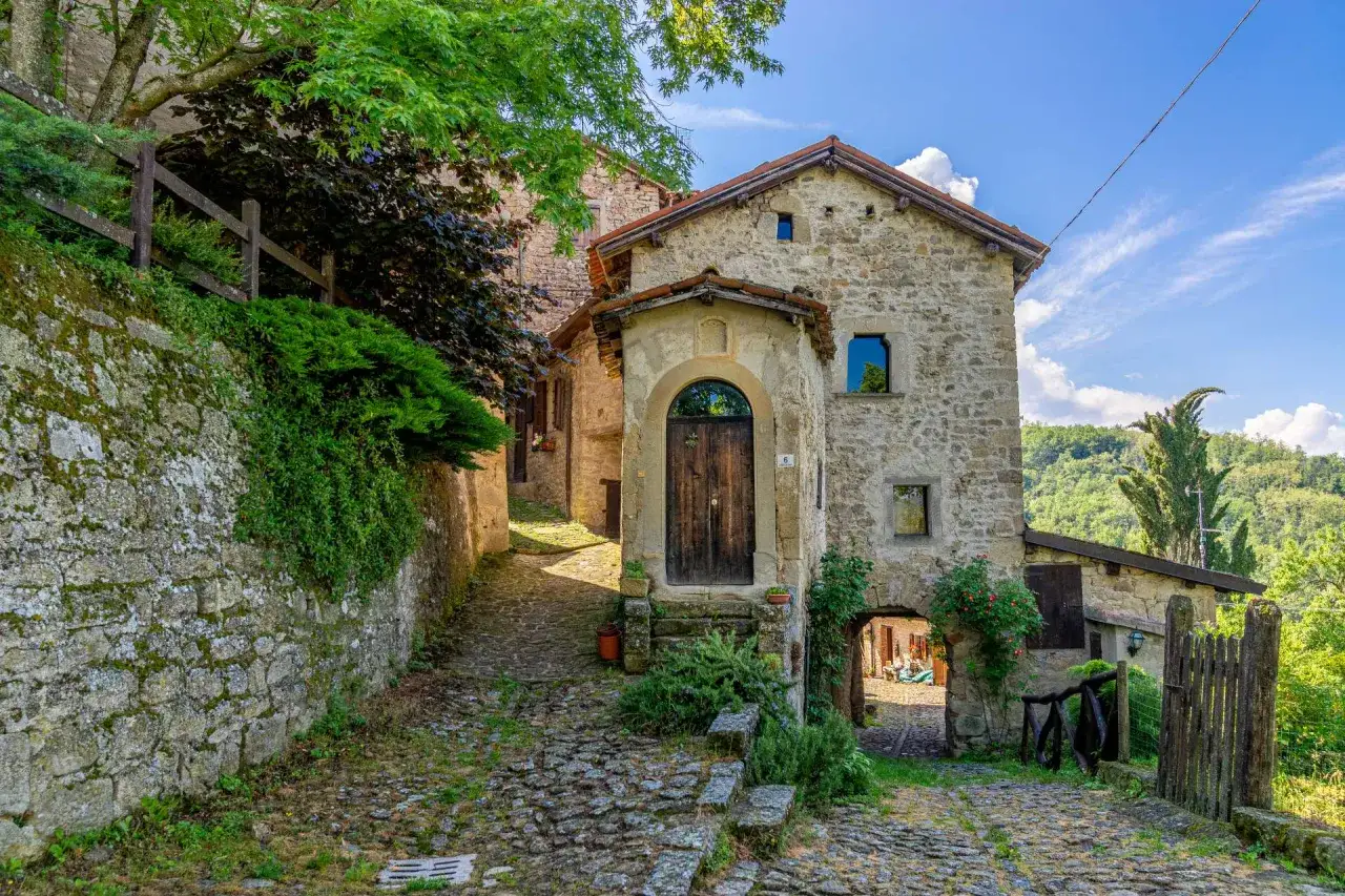 Antica via acciottolata in un borgo di pietra, con un arco che conduce a un cortile e un'antica casa che ricorda la scola.