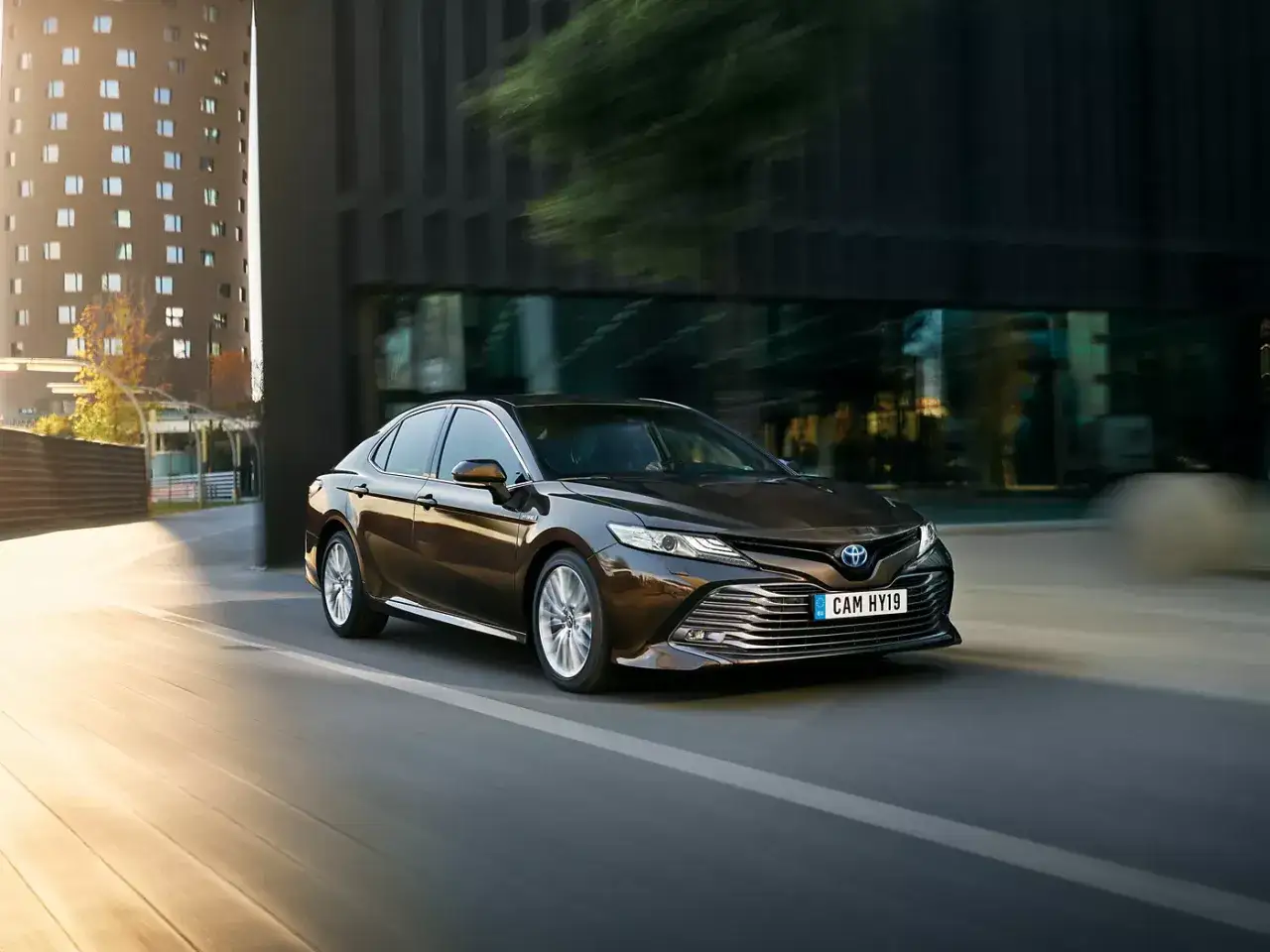 Brun Toyota Camry Hybrid k&ouml;rs genom en stadsmilj&ouml;. En perfekt begagnad bil f&ouml;r dig som s&ouml;ker komfort och stil.