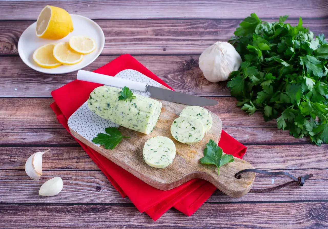 Un rouleau de beurre maître d'hôtel, garni de persil frais, est prêt à être découpé. Citrons, ail et persil complètent la scène.