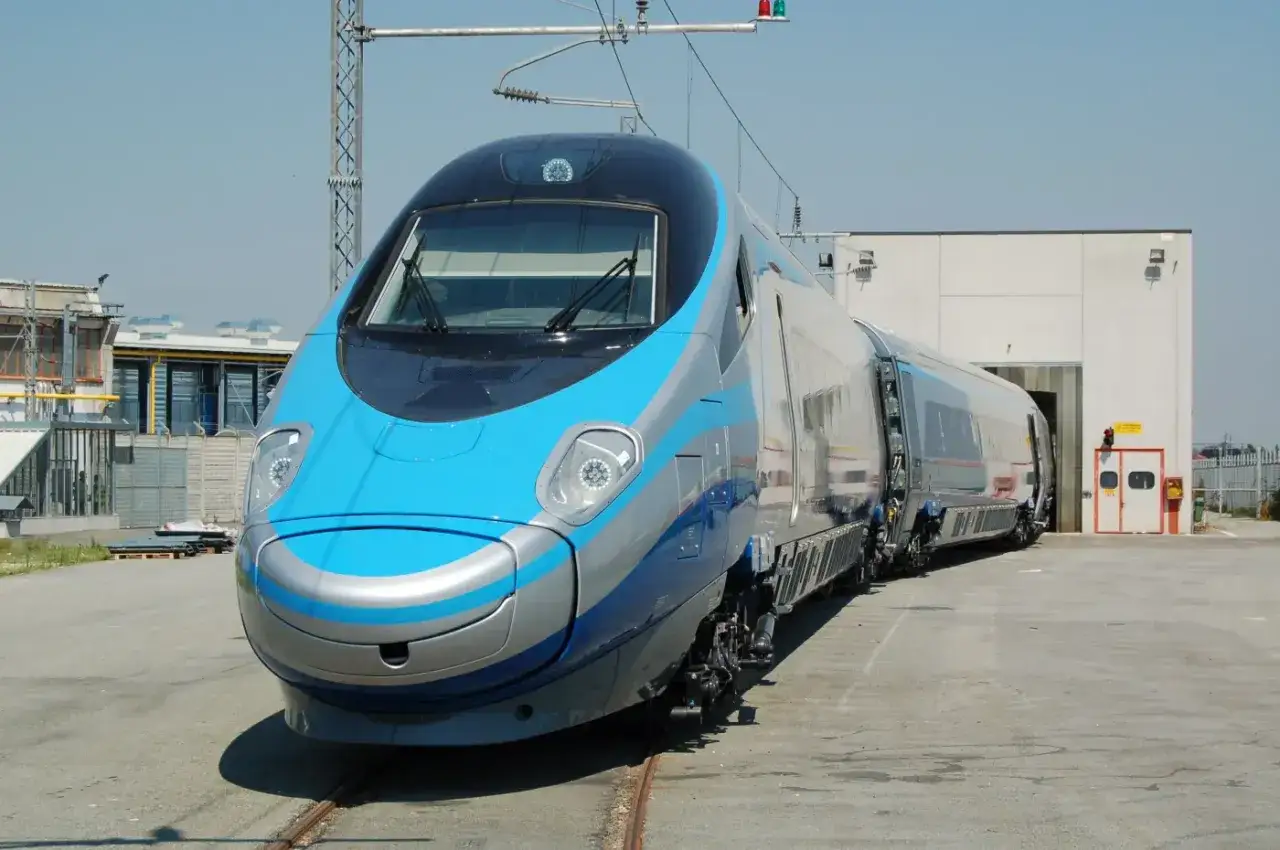 Pendolino w całej okazałości, gotowe do osiągnięcia zawrotnej prędkości. Błękitno-srebrny pociąg stoi na torach przed halą.
