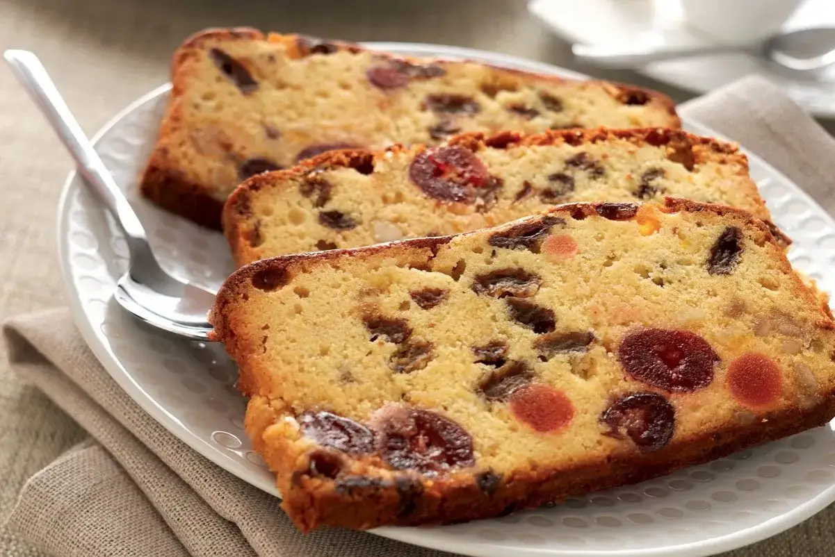 Trois tranches de cake aux fruits confits, parsem&eacute; de raisins secs et de cerises, sur une assiette blanche. La meilleure recette de cake aux fruits confits.