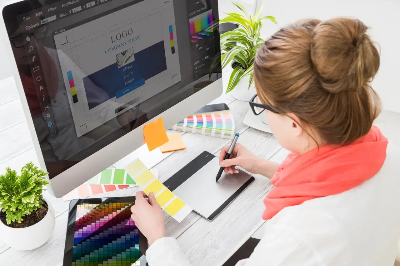 Frau im Grafikdesign-Studium arbeitet am Computer und mit Grafiktablett. Farbpaletten und Design-Software sind sichtbar.