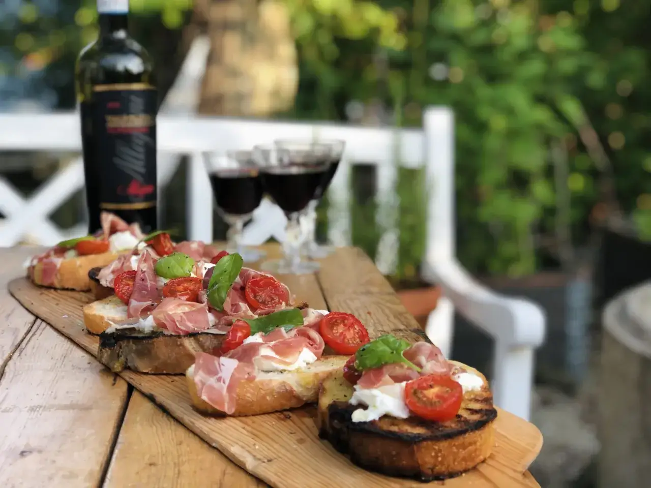 Bruschetta med mozzarella, prosciutto och tomater på en träbricka, med vin och glas i bakgrunden. En perfekt sommardessert.