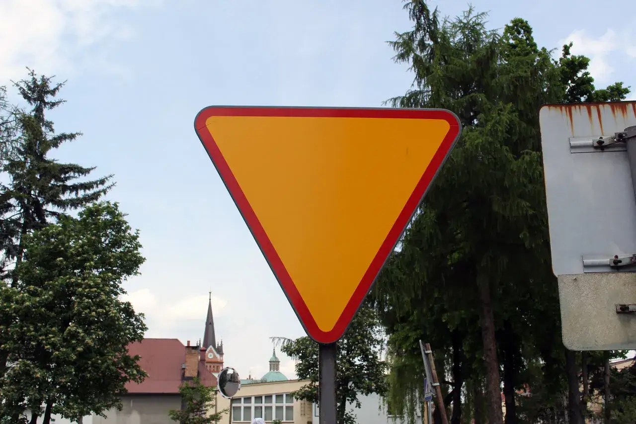 Żółty trójkąt z czerwoną obwódką, czyli znak ustąp pierwszeństwa, stoi na tle drzew i nieba.