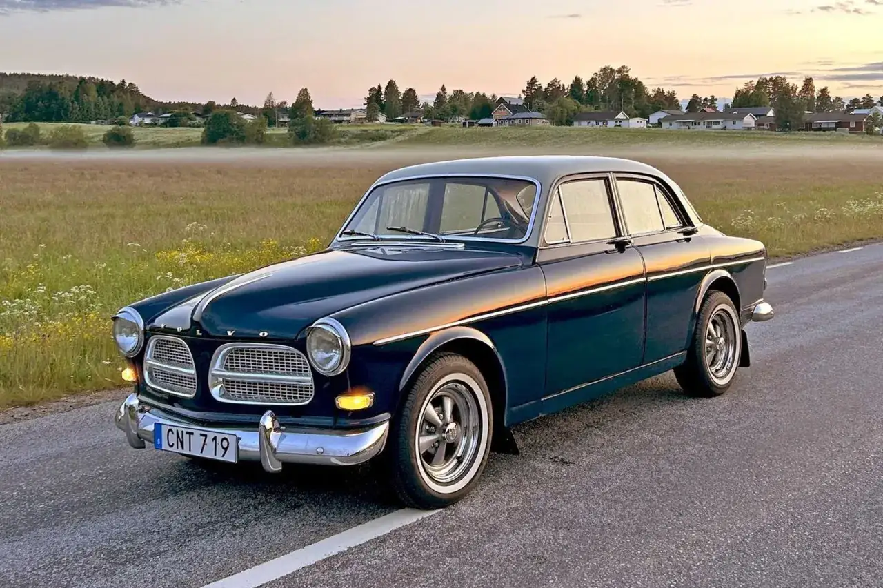 En mörkblå Volvo Amazon-bil står parkerad vid en väg i solnedgången.