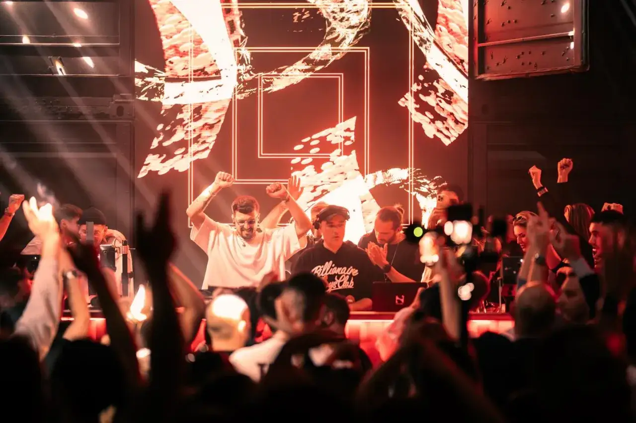 Un dj spagnolo famoso fa impazzire la folla con le braccia alzate. Luci rosse e uno schermo gigante creano un'atmosfera elettrica.