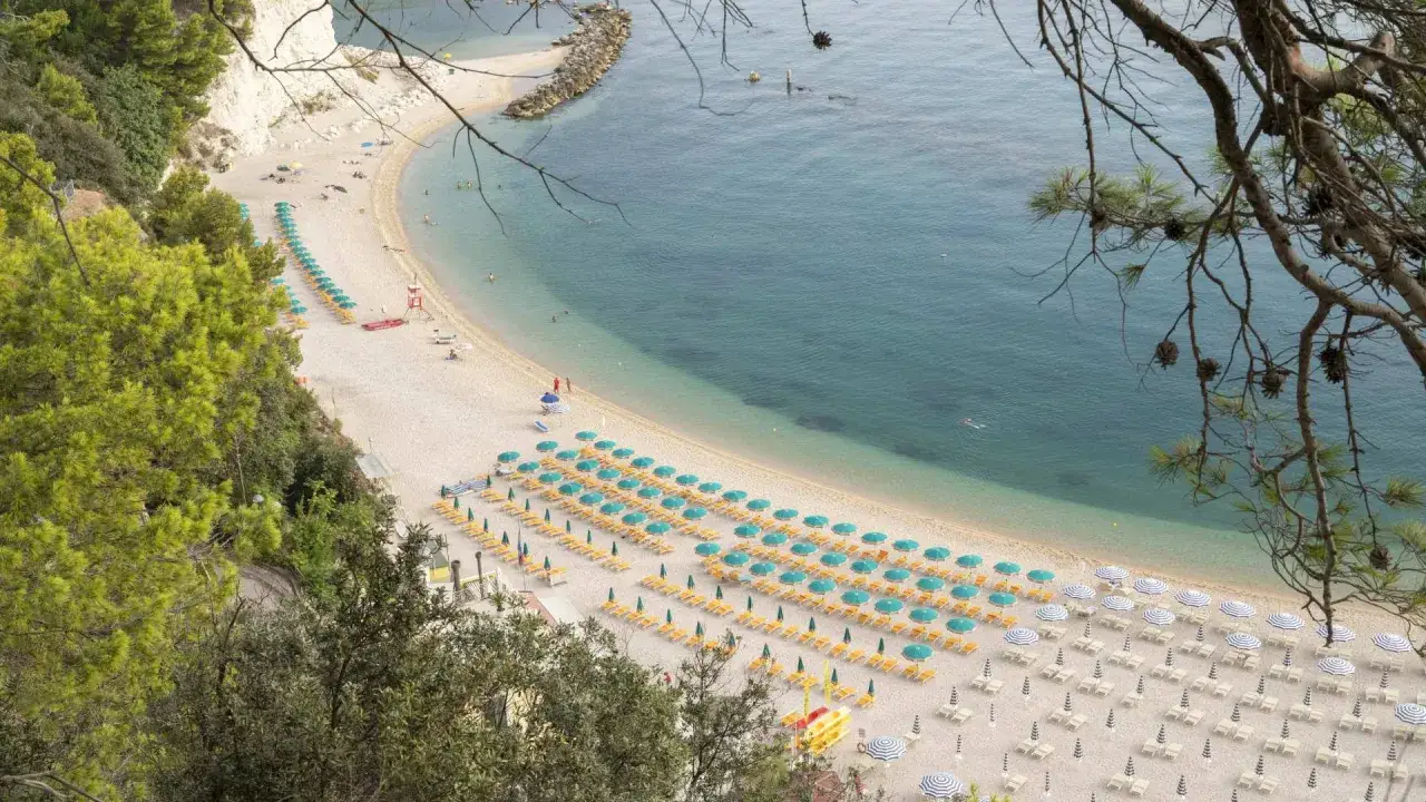 Spiaggia di Sirolo, Marche, con file ordinate di lettini e ombrelloni verdi e gialli che si estendono lungo la costa sabbiosa bagnata da un mare azzurro e limpido.