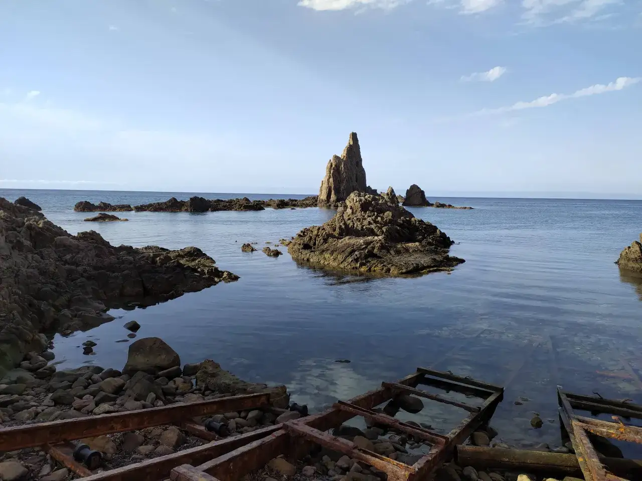 Rocas escarpadas emergen del mar azul, evocando el mítico arrecife de las sirenas. El agua cristalina revela el lecho rocoso y restos oxidados de estructuras antiguas.