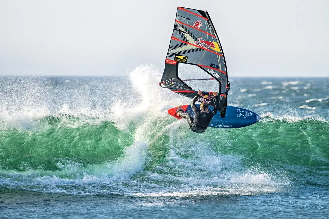 Liam Dunkerbeck fliegt mit seinem Windsurfbrett über eine grüne Welle, die Gischt spritzt.