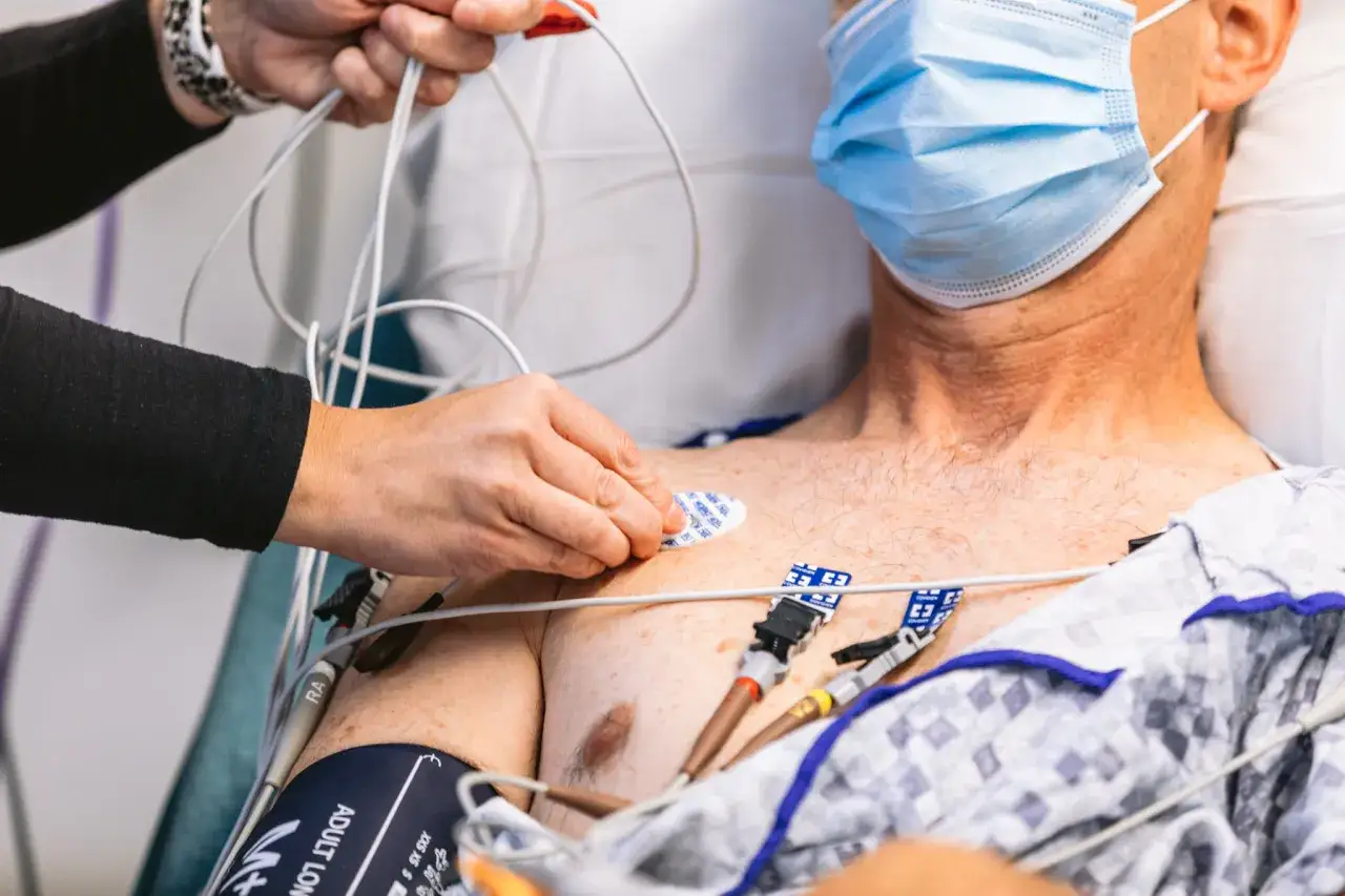 Pielęgniarka przykłada elektrodę do klatki piersiowej pacjenta, przygotowując **ekg serca**. Widać przewody aparatu i maskę na twarzy mężczyzny.