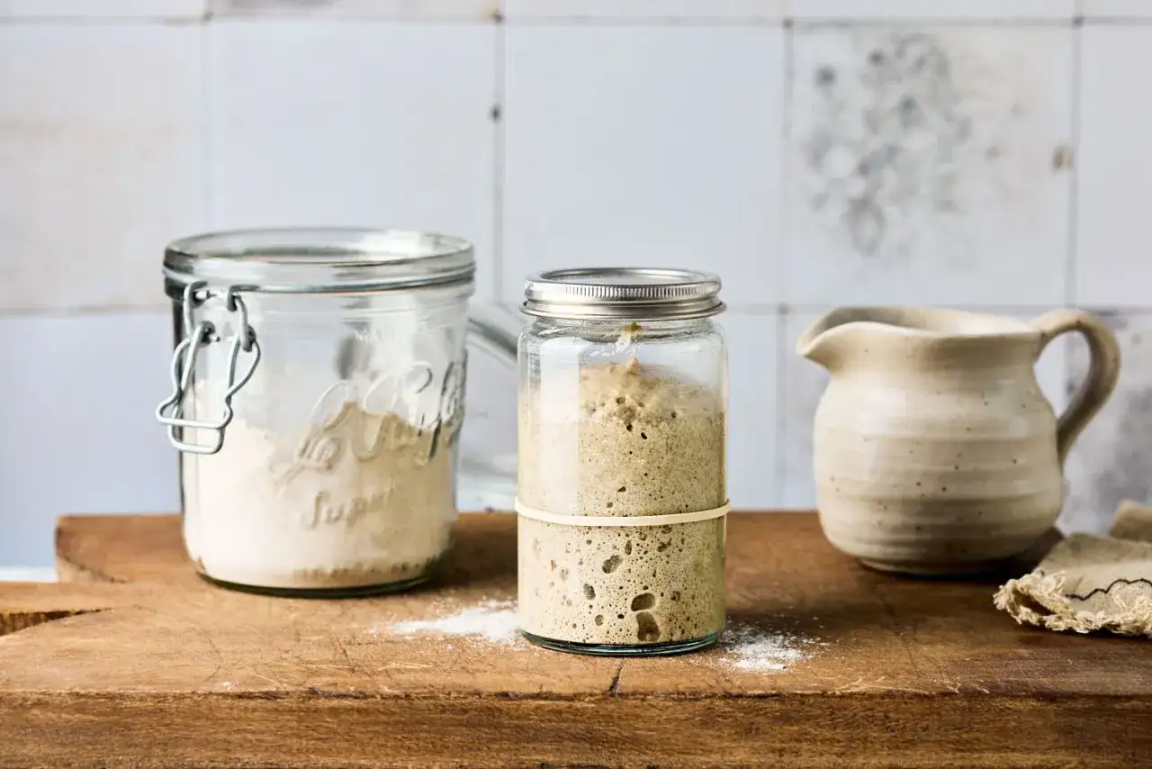 Zwei Gläser mit Sauerteig: eines mit trockenem Mehl, das andere mit aktivem, sprudelndem Anstellgut. Ein kleiner Krug und ein Tuch stehen daneben.