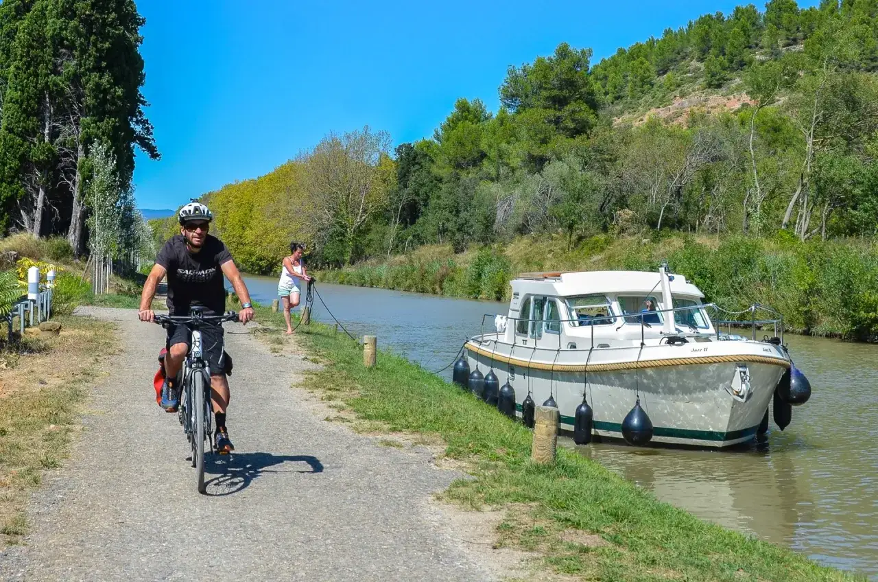 Ciclista pedalea por el camino del Canal du Midi, junto a una barcaza y una mujer que sujeta una cuerda.