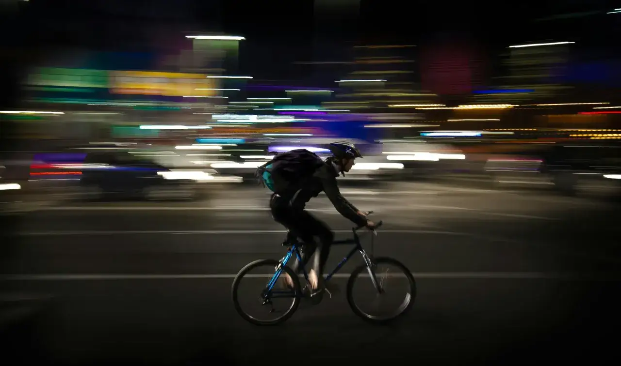 Rowerzysta w kasku mija rozmazane światła miasta. Z tyłu roweru miga czerwone światło pozycyjne, zapewniając widoczność.
