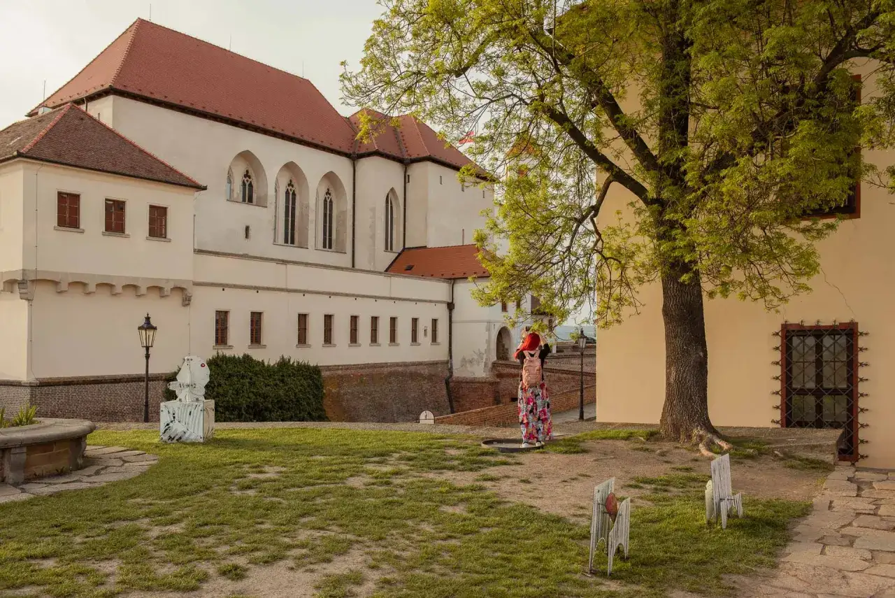 Kobieta w kolorowej sukience podziwia zamek w Brnie. W oddali widać zabytkowe budowle i zielony dziedziniec.