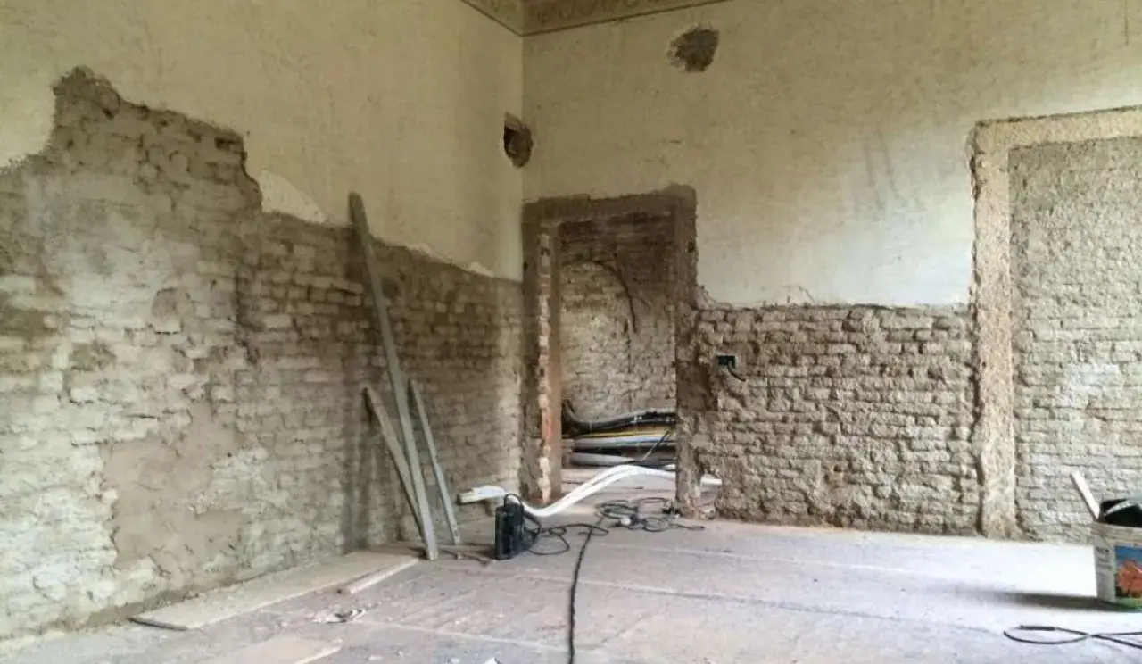 Interno in fase di ristrutturazione, con un muro in pietra e intonaco parzialmente rimosso, travi di legno e cavi elettrici a vista.
