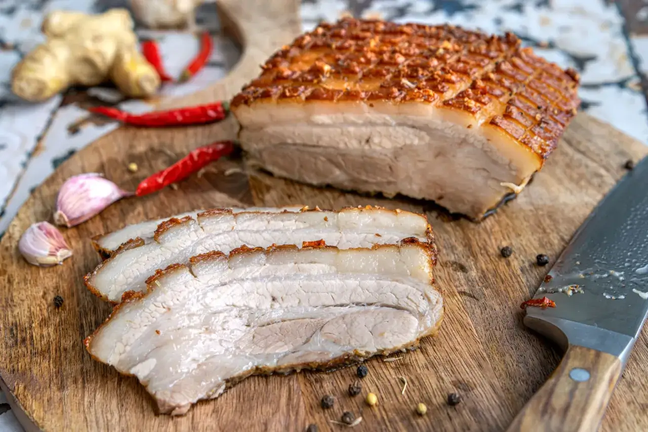 Ein butterzarter Schweinebauch mit knuspriger Kruste, daneben geschnitten, mit Chili, Knoblauch und Ingwer auf einem Holzbrett.