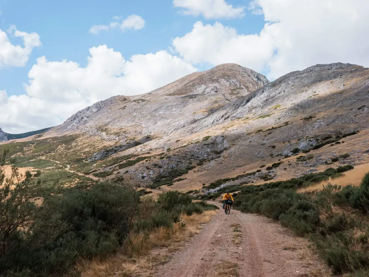 Ciclista en un camino de tierra entre matorrales, explorando rutas bikepacking España bajo un cielo azul con nubes.