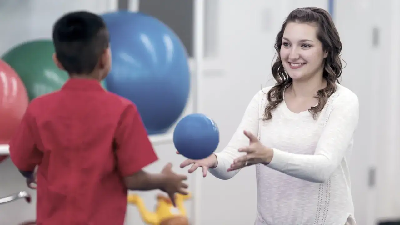 Ni&ntilde;o y terapeuta juegan con una pelota azul. La terapeuta sonr&iacute;e, ayudando al ni&ntilde;o a entender la dispraxia que es y c&oacute;mo superarla.