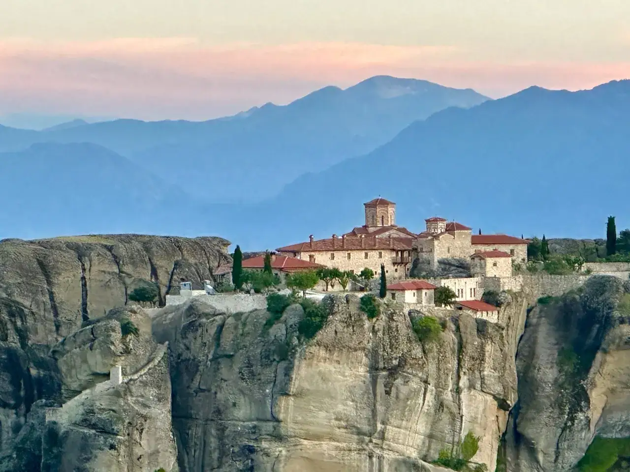 Meteora Kloster thront auf steilen Felsen, umgeben von blauen Bergketten im sanften Abendlicht.