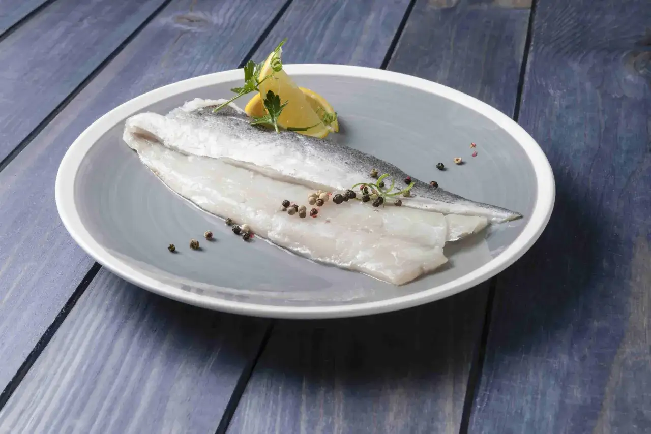Filete de pescado blanco crudo, sazonado con granos de pimienta y adornado con limón y perejil, listo para cocinar.