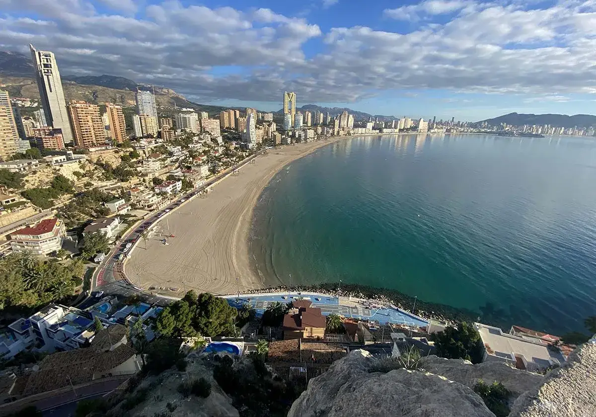 Vista panor&aacute;mica de la **mejor playa de Benidorm, Levante o Poniente**, con su extensa arena dorada, el mar turquesa y la silueta de los rascacielos.