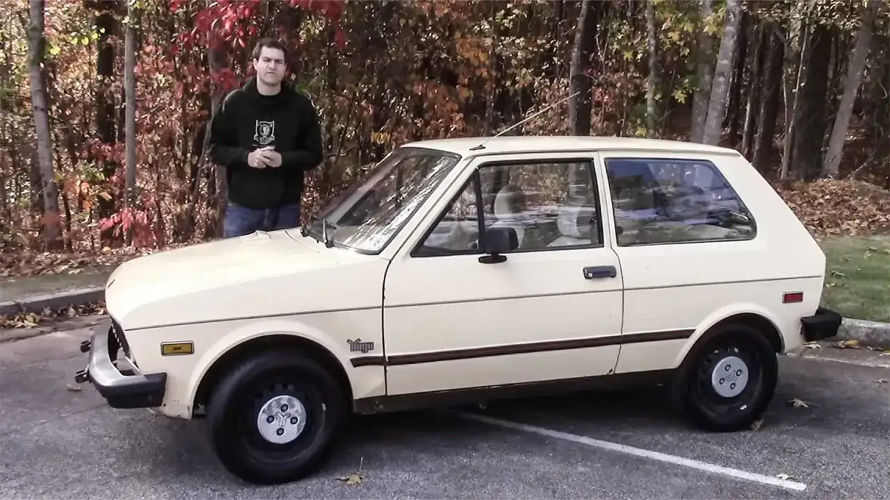 En man står bredvid en beige yugo bil. Bilen har en klassisk design och står parkerad utomhus bland höstträd.