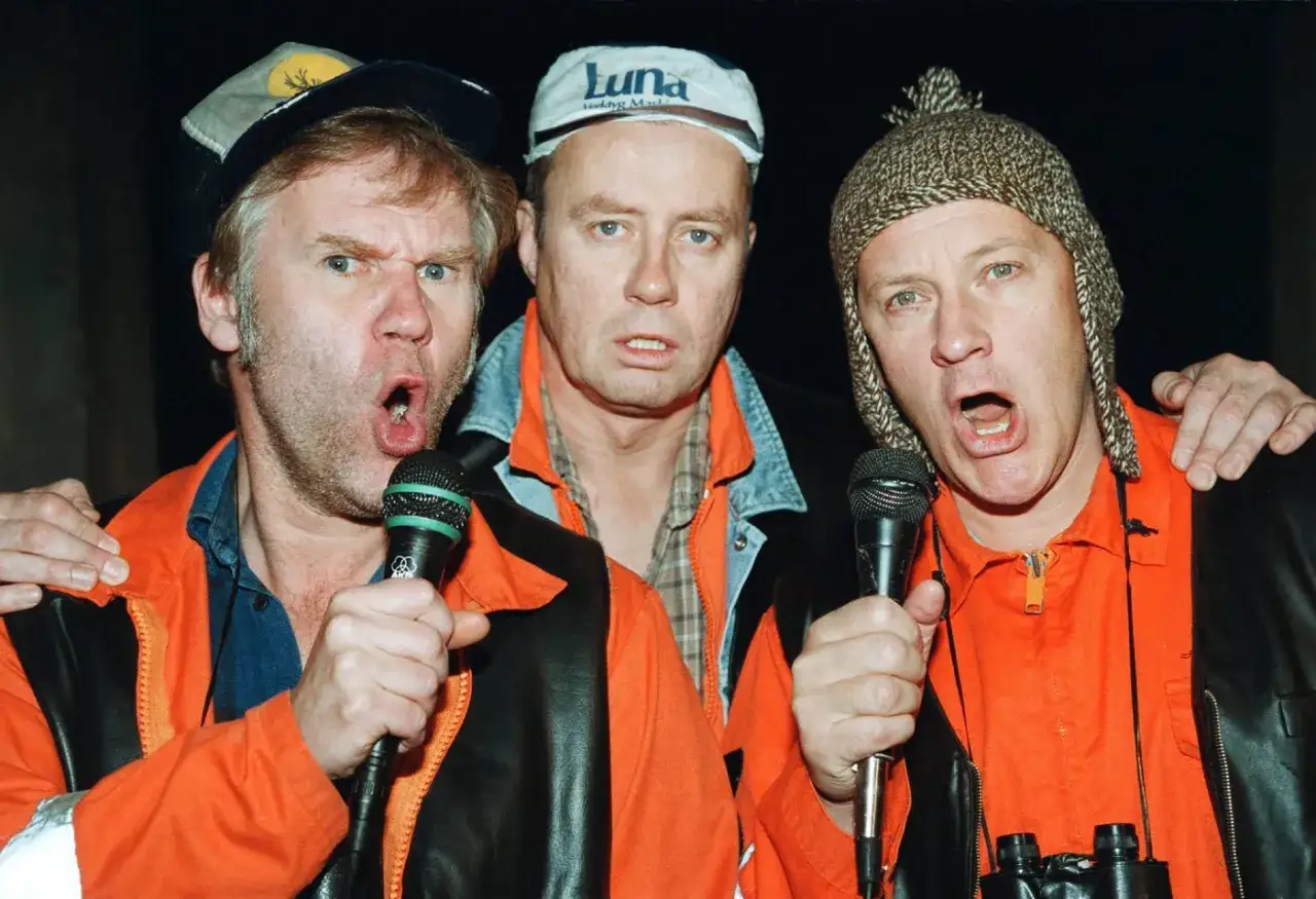 Tre män med mikrofoner, klädda i orangea overaller och mössor. Kanske är det rollistan i Pistvakt som sjunger?