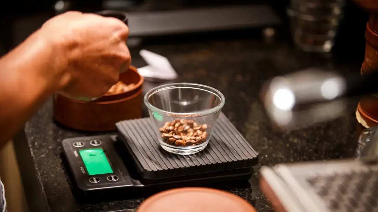 Eine Hand mahlt Kaffee. Die genaue **Kaffee kochen Menge** wird auf einer Waage mit einer kleinen Schale voller Bohnen abgewogen.