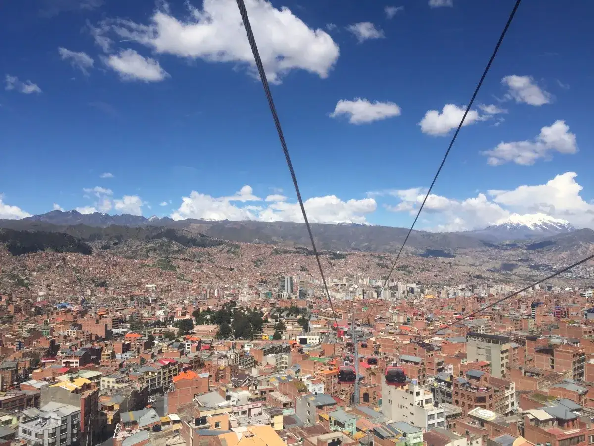 View from a la paz gondola over a sprawling city with mountains in the distance under a blue, cloudy sky.