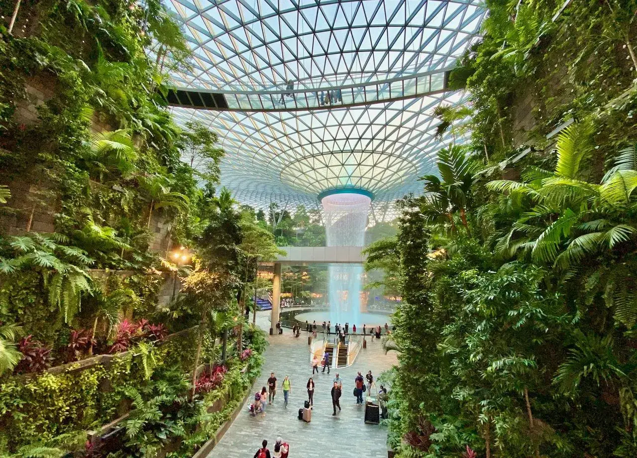 Imposanter Wasserfall im tropischen Garten des **Singapore Flughafens** unter einem Glasdach. Menschen spazieren durch das grüne Paradies.