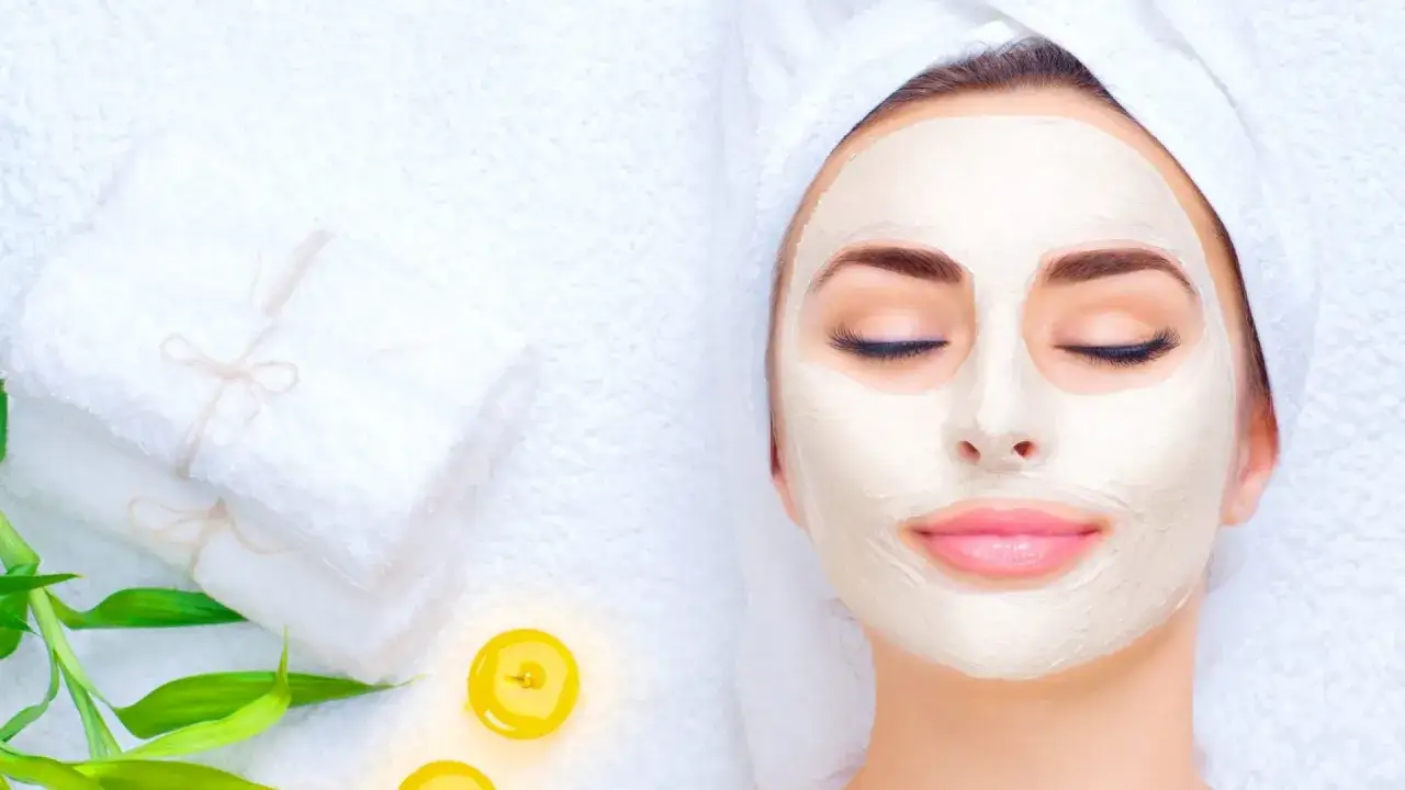 Entspannende Gesichtsmaske morgens oder abends. Eine Frau mit geschlossenen Augen und einer cremigen Maske im Gesicht, umgeben von Handt&uuml;chern und Kerzen.