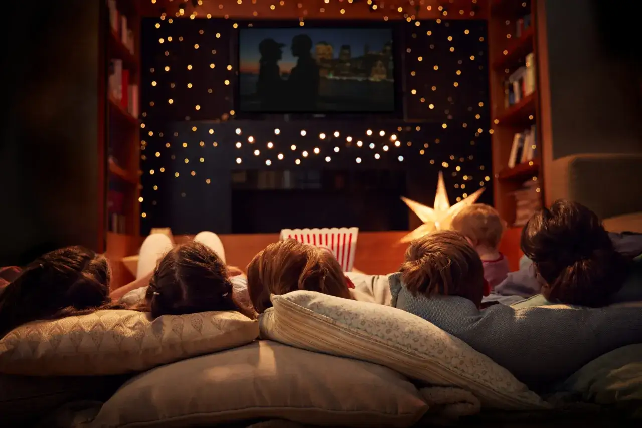 Bambini accoccolati su cuscini, guardano un film con luci natalizie e popcorn. Una serata perfetta per vedere un film ragazzi.