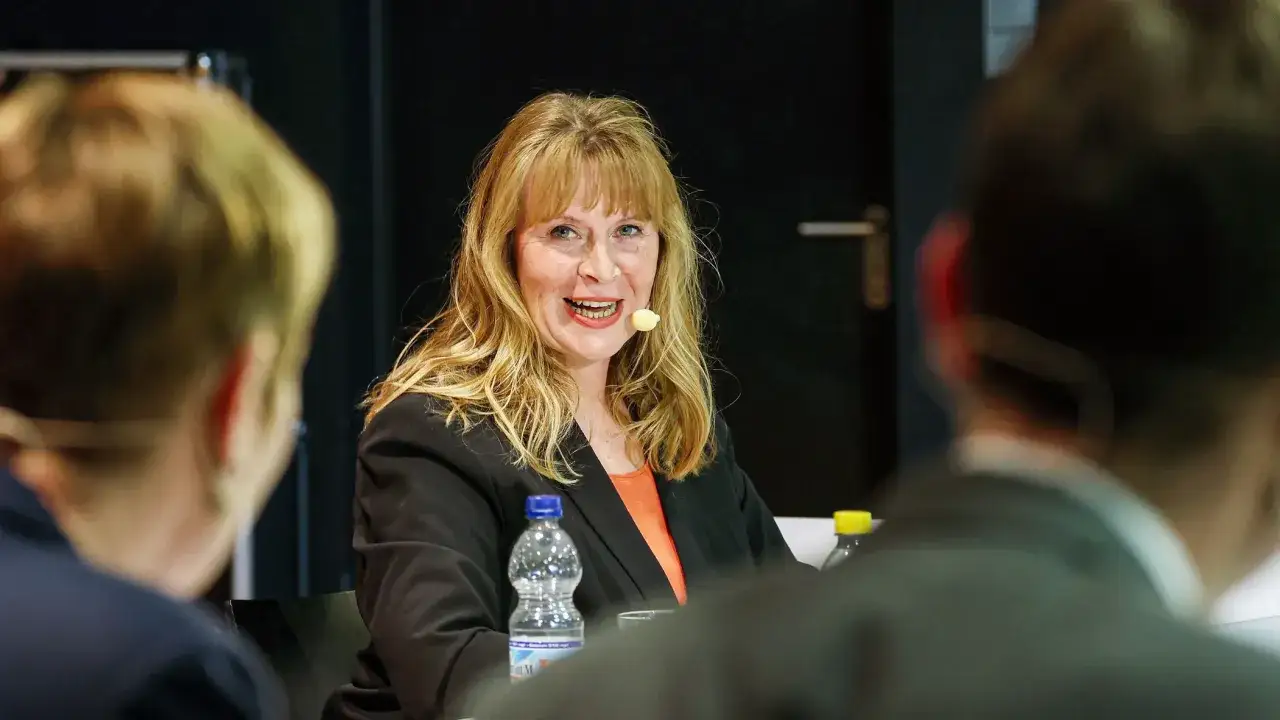 Petra Weber, lächelnd, mit Mikrofon, sitzt im Gespräch mit anderen Personen.