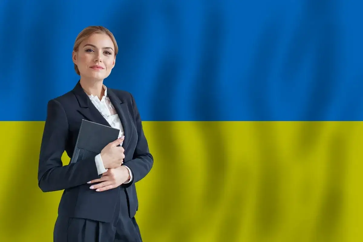 Uśmiechnięta kobieta w garniturze, trzymająca teczkę, na tle flagi Ukrainy. Symbolizuje zatrudnienie obywatela Ukrainy.