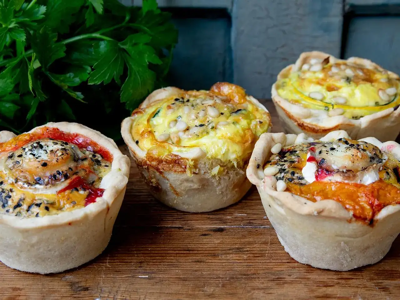 Mini Quiche Rezept: Vier herzhafte T&ouml;rtchen mit Eierf&uuml;llung, Pinienkernen und Kr&auml;utern auf Holzuntergrund.
