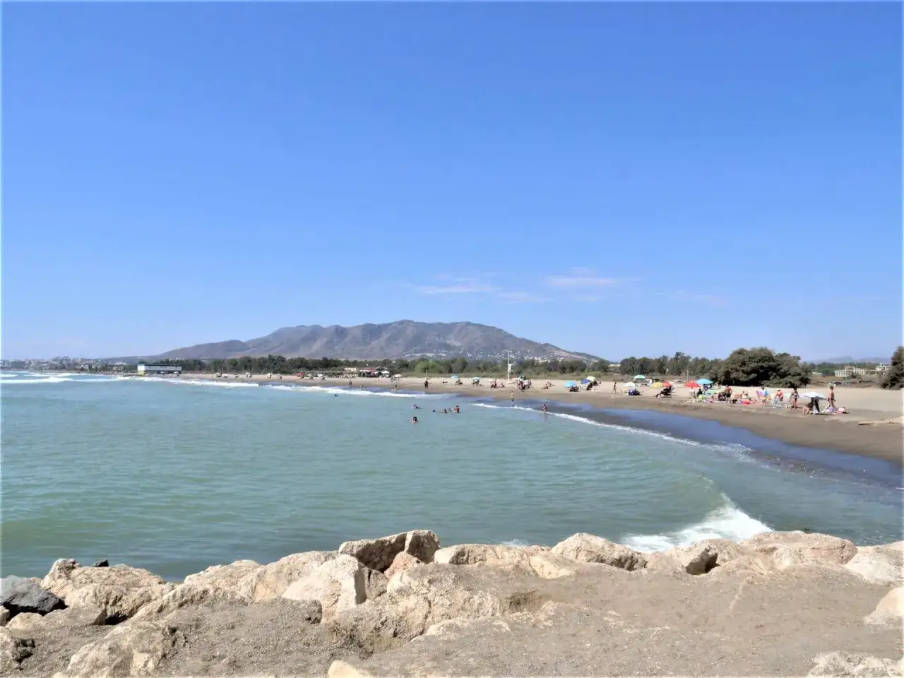 Słoneczne malaga plaże z ludźmi kąpiącymi się w morzu i odpoczywającymi na piasku, w tle góry.