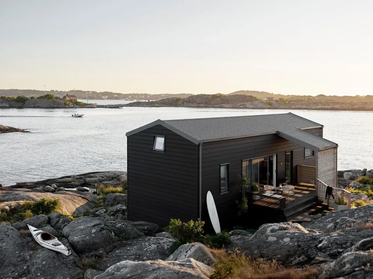 Ett modernt, mörkt grått hus med svarta knutar står på en klippig kust. En kajak ligger på stenarna vid vattnet.