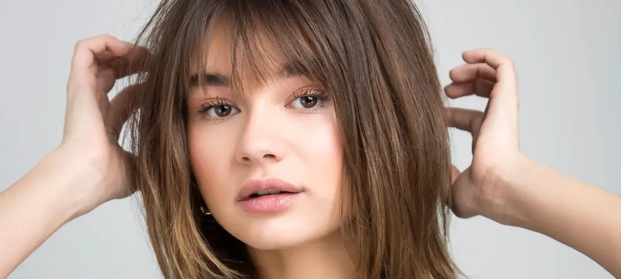 Chica con cabello castaño y flequillo, ideal para un corte de pelo cara redonda. Sus manos juegan con su melena.